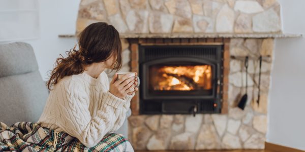 Young woman sitting at home by the fireplace with a hot tea or coffee mug and warming her hands, she is wearing white woollen sweater. Cold houses in Europe concept during energy and gas crisis.